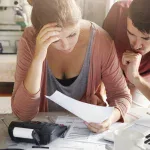 Un couple inquiet examinant des documents financiers et des factures sur une table avec une calculatrice et un ordinateur portable.